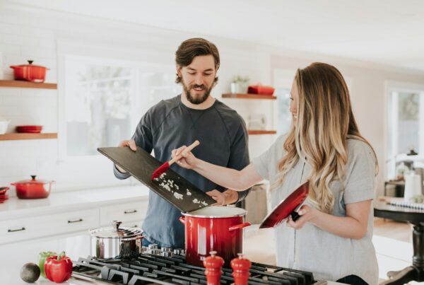 A couple cooking together in a well-lit kitchen, smiling as they use red pots and pans. The kitchen features red accents throughout, adding a warm and vibrant touch.